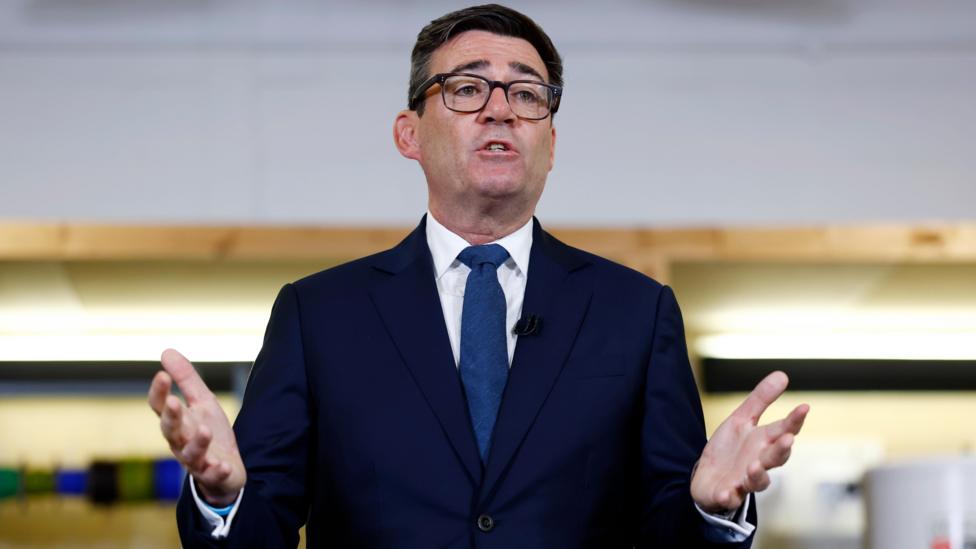Andy Burnham wears a dark suit and blue tie and is mid speech, gesturing forwards with his hands
