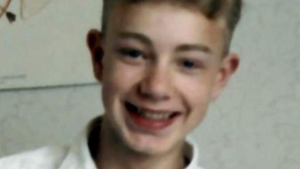 Close-up photograph of Alan Watson with short hair wearing a light-coloured collared top, looking towards the camera against a plain indoor background.