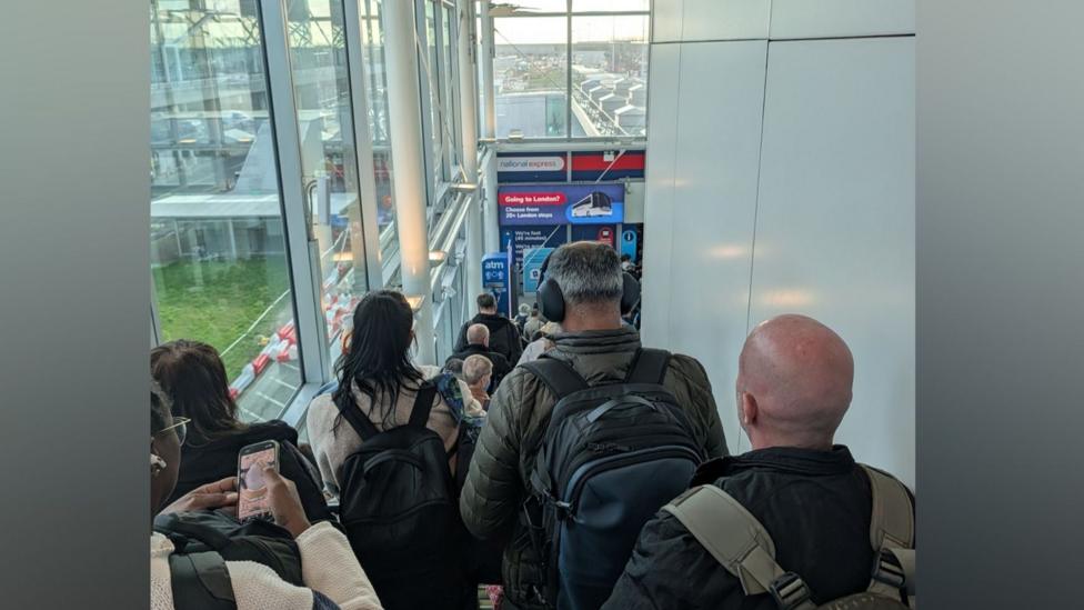 People in a long queue backing up some stairs at Stansted Airport. The individuals are waiting and in front of them are glass panels that make up the walls of the airport terminal.