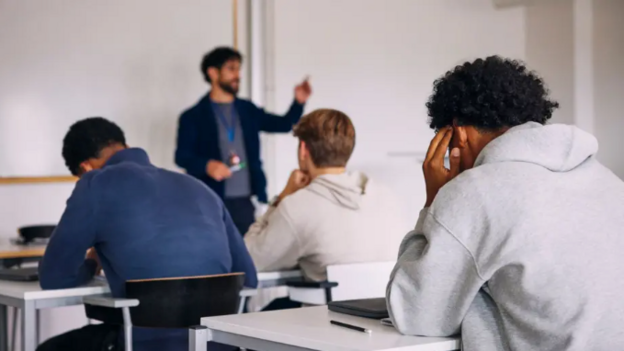 A male teacher stands in front of a white board raising his hand and addressing a class. He has dark hair and a beard and wears a grey t shirt, a blue jacket and a lanyard. In front of him, with their backs to the camera are three male students at desks. They wear cream, blue and grey jumpers.