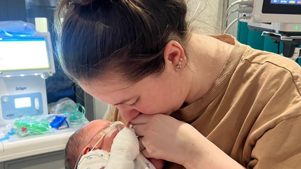 Sophie holding her newborn daughter Florence. Tubes can be seen attached to Florence's face. Her left arm is bandaged. Sophie has brown hair which is tied back. She wears a beige t-shirt. Her eyes are closed and she is not looking at the camera.