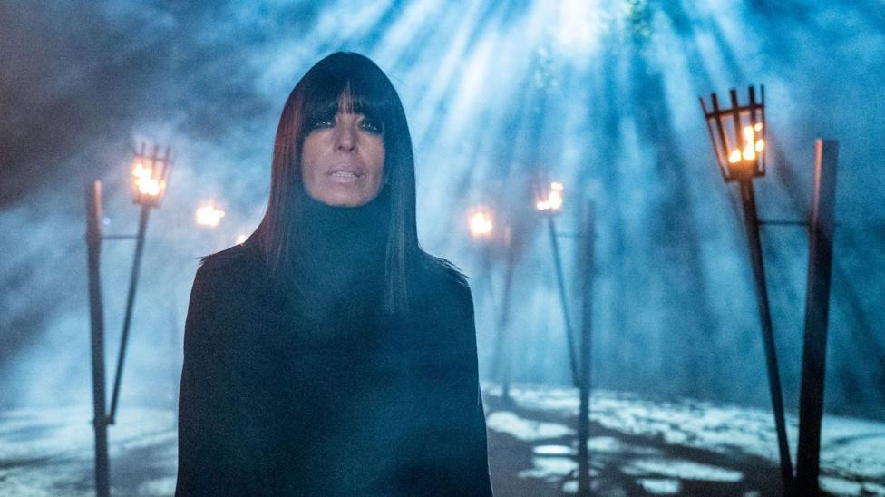 Claudia Winkleman in the Traitors. She is bathed in rays of moonlight poking through a forest canopy as braziers light the path behind her