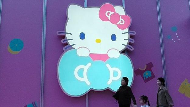 Три женщины проходят мимо большой светящейся вывески Hello Kitty на фиолетовой стене магазина в Шанхае, Китай