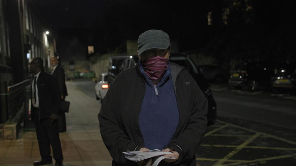 It is nighttime and Jessica is wearing a cap and a red scarf across her face. She is standing on the pavement holding a bunch of leaflets.
