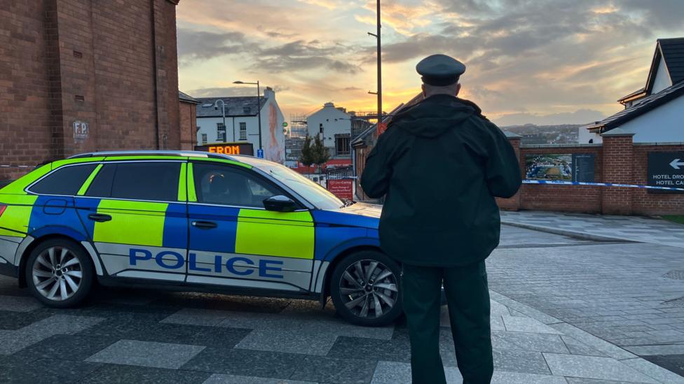 A police officer stands with his back to the camera. He is in uniform and standing beside a police car. Police tape can be seen in front of buildings in the background.