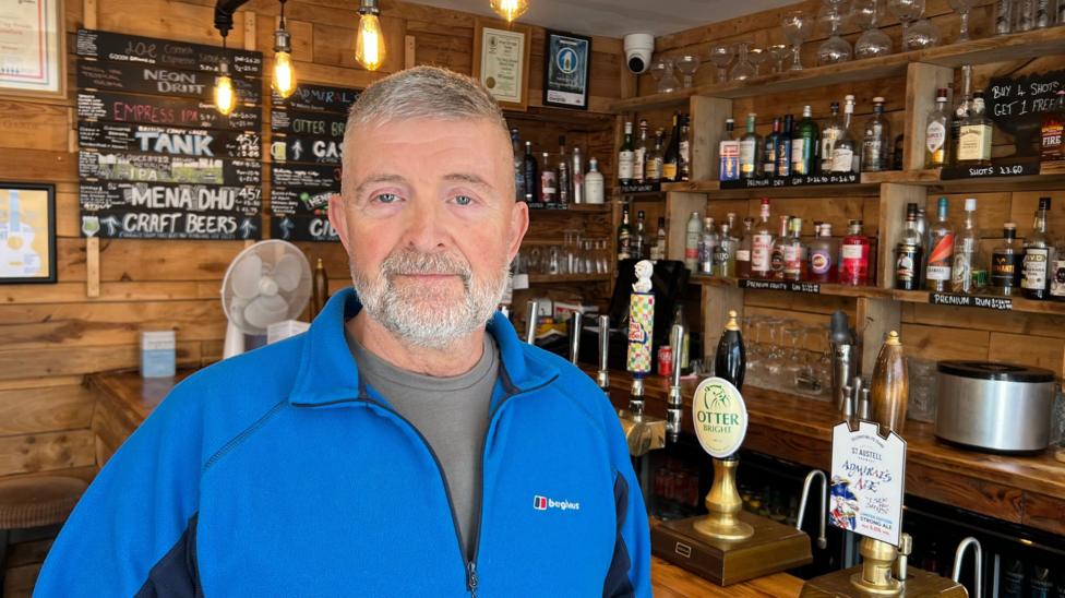 A man in a blue top standing in a wooden bar area in a pub.