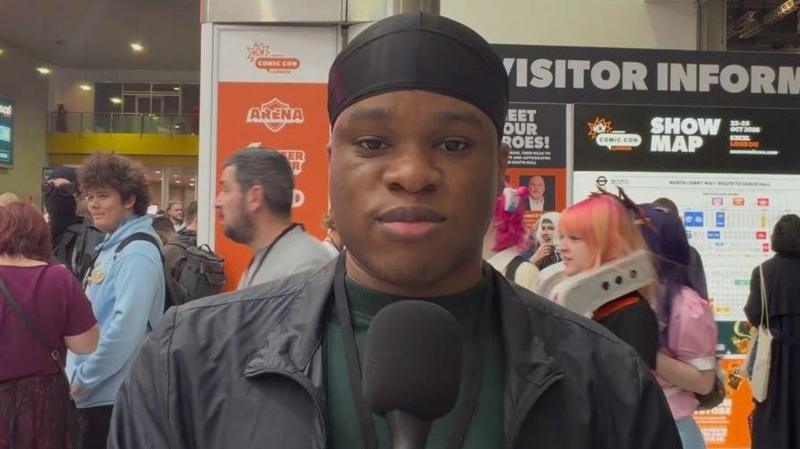 Itanna Alakija kameraga qarab turgan holda tabassum qilmoqda, orqasida Comic Con tashrif buyuruvchilari aylanib yurishadi. U sochiga qora himoya qopqog‘i taqib olgan, yashil sviter va qora kurtka kiygan.