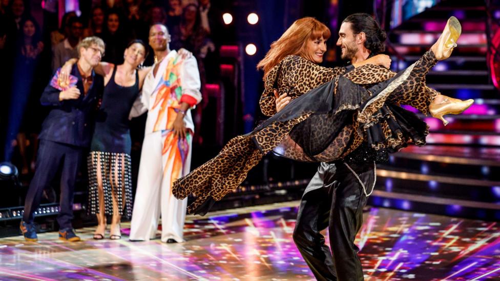 Ведущие Strictly на заднем плане обнимают друг друга за плечи, а Сару Стори несёт танцевальный партнёр Джулиан Кайон