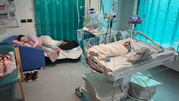 Inside the paediatric ward at Grange University Hospital in Cwmbran. Sophie Price is a young woman who is seen lying on her side on top of a sofa bed. Her daughter Florence is inside a hospital baby crib. A breathing tube can be seen over her nose. She is wrapped in a white blanket.