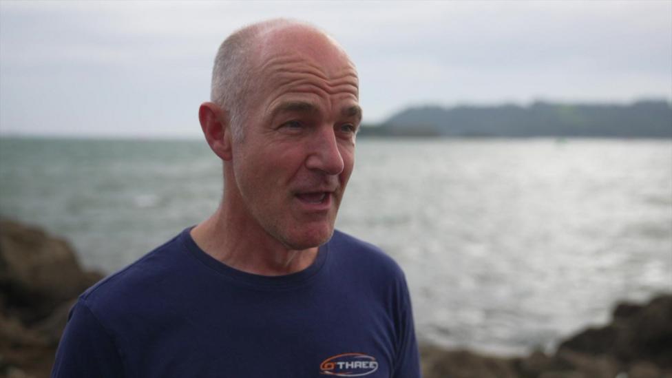 Dominic Robinson - a diver wearing a navy blue t-shirt speaks to the camera with the sea in the background.