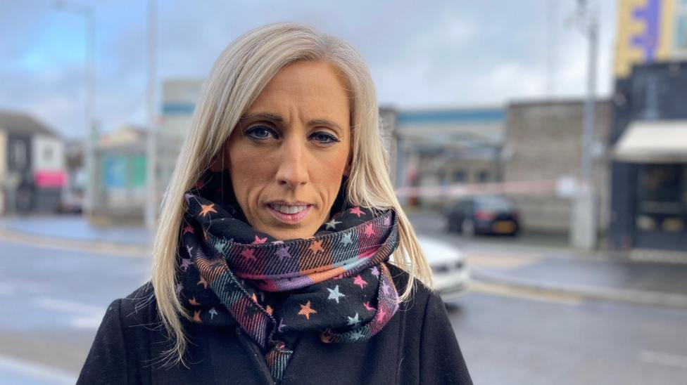 Karla Lokhart Lurgan politsiya bölimi oldida turibdi. U uzun sarğiş soçli örtaça yoşdagi ayol bölib, rang-barang, yulduz naqşli römol va qora palto kiygan. U betaraf yuz ifodasi bilan kameraga qarayapti.