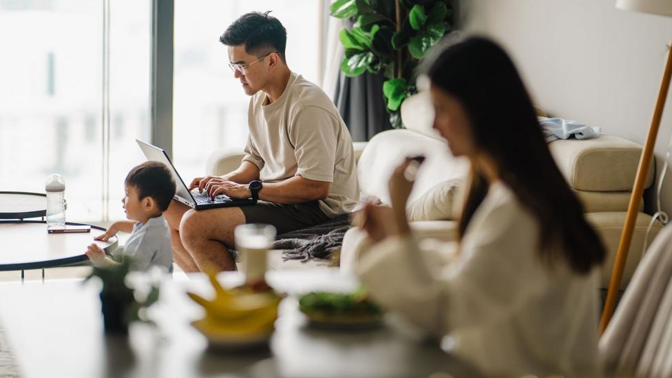 Stock photo shows couple loking at a computer and eating in a living room with a toddler in view.