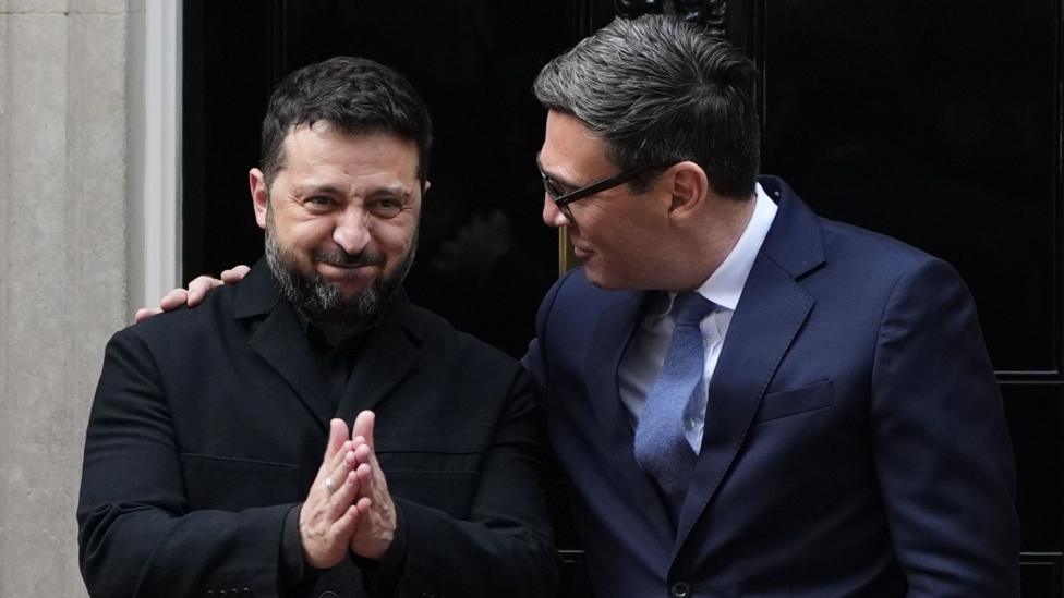 Volodymyr Zelensky smiling with his hands together standing next to Andy Burnham who has his arm around him.