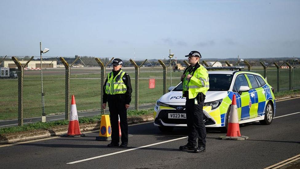 Two police officers stand with a police car outside RAF Fairford