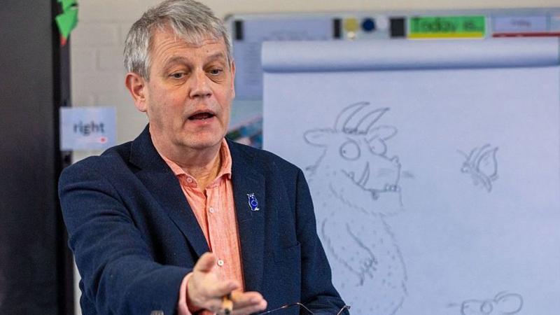 Аксель Шеффлер, мужчина с седыми волосами, в тёмно-синей куртке поверх оранжевой рубашки, жестикулирует во время выступления перед белой доской с наброском Груффало.
