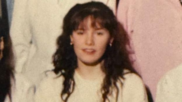 Cropped group photograph showing Pauline with long curly hair wearing a light-coloured top, looking towards the camera, with other partially visible people standing nearby.