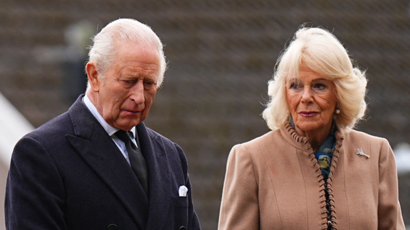 King Charles and Queen Camilla stand outside. Charles looks down with a serious expression, wearing a dark coat. Camilla looks to the side in a tan coat.