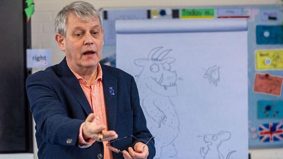 Мужчина с седыми волосами, в персиковой рубашке и синей куртке, держит очки и стоит перед белой доской для рисования.
