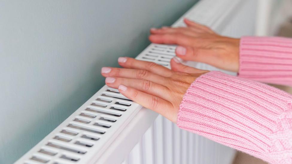 Ikkala qo‘li bilan radiatorning yuqori qismiga tegayotgan ayol. U uzoq yengli pushti trikotaj sviter kiygan va tirnoqlari estetik tarzda parvarishlangan.