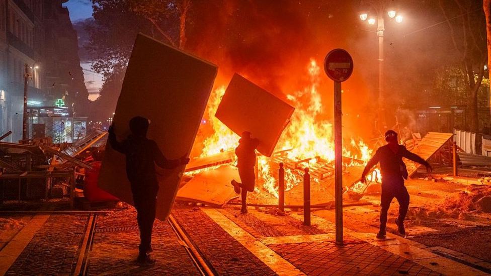 Протестующие поджигают баррикады в Марселе ночью. На городской улице видно большое пламя. Два человека идут к нему с большими прямоугольными щитами, предположительно, чтобы подбросить в огонь. Третий человек идёт рядом с огнём. В кадре видны фонарные столбы, столбики и деревья, а слева — ряд магазинов.
