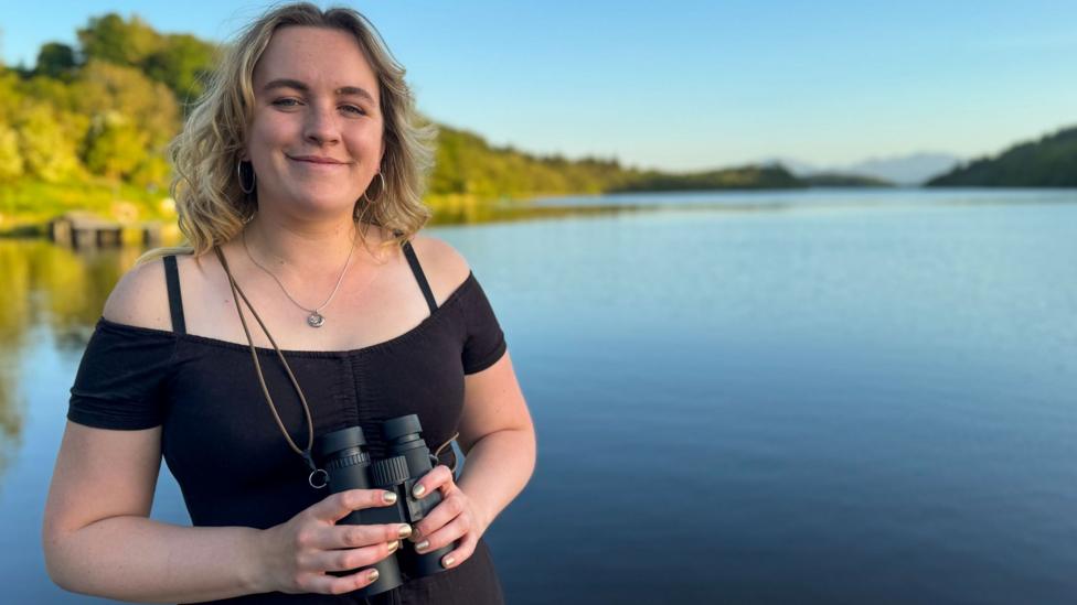 Caitlin Andrew is a young woman with shoulder length blonde hair. She is smiling as looks into the camera while holding a pair of black binoculars. Behind her is a loch with trees along its shores.