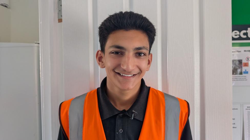 Param is pictured wearing a high-visibility orange safety vest over a black collared shirt, standing indoors in front of a white panelled door. Part of a noticeboard and a white appliance are visible in the background.