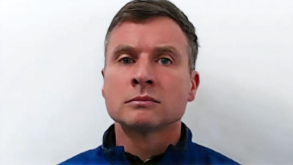 Head-and-shoulders portrait of a person wearing a blue zip-up jacket against a plain light-colored background, facing the camera with a neutral expression.