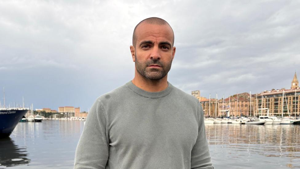 Arnaud Blaise poses for a photograph at a port in Marseille during the day. He has a serious expression, short black hair and facial hair. He is wearing a grey crew-neck jumper. Behind him is a calm marina with numerous white boats docked along the right hand side. It is an overcast day.