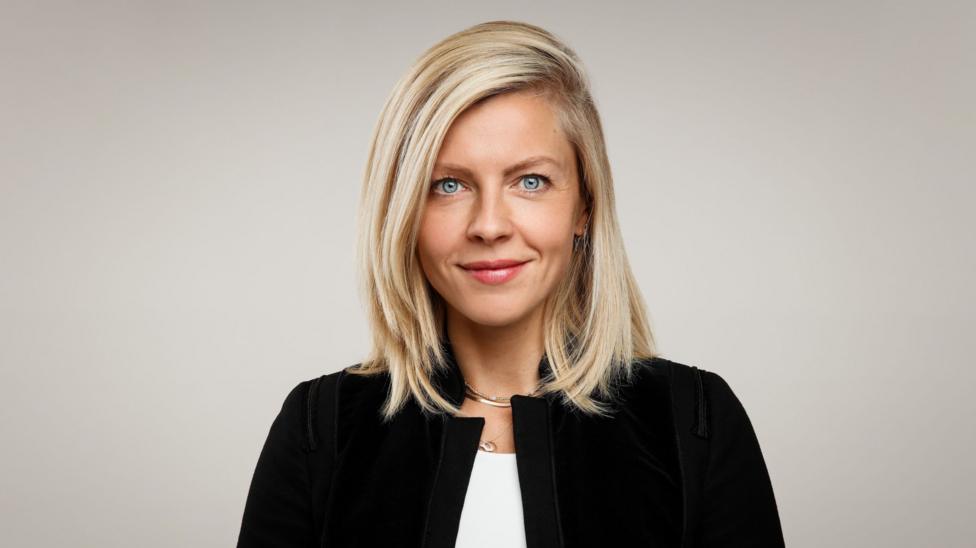 Deirdre O'Neill looks at the camera and smiles. She has blonde hair, blue eyes, and is wearing a black jacket and white blouse. She's standing against a plain grey backdrop.