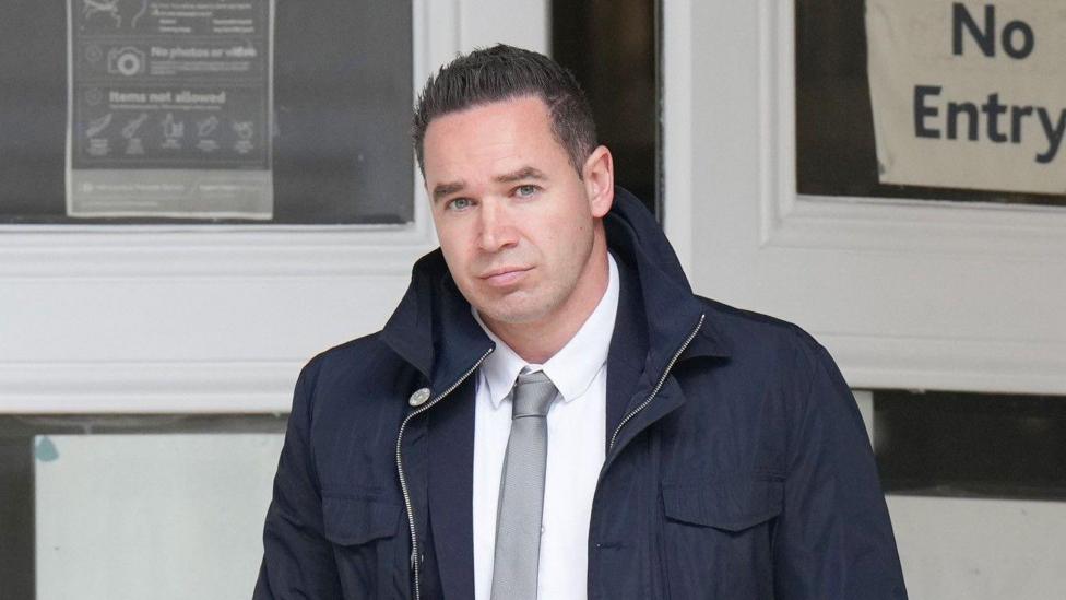 A man with short black hair wearing a large dark blue coat. He is looking into and walking towards the camera.