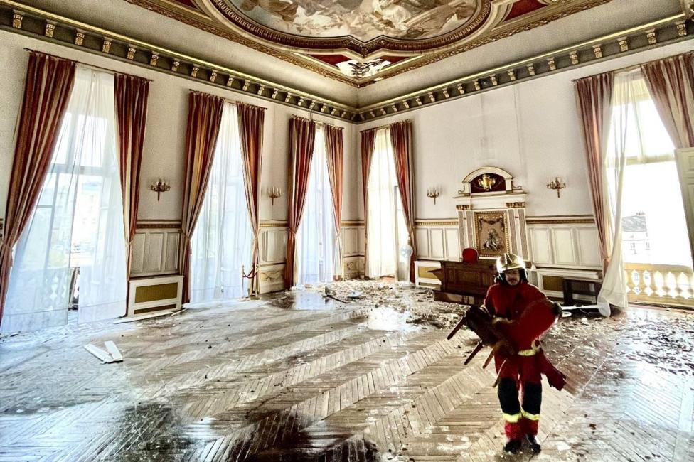 An ornate, large room in Limoges city hall. The parquet floors are covered in rubble and a fireman is carrying a chair across the room. The ceiling has a scene of a blue sky with angels and divinities painted on it.