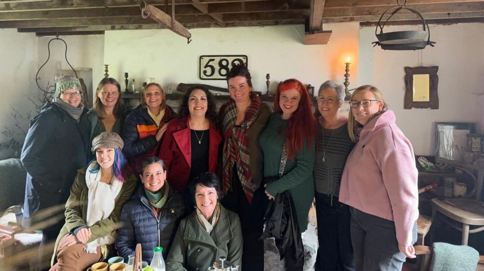 A group of 11 women, all wearing colourful jumpers, standing together in a cottage environment smiling widely at the camera.