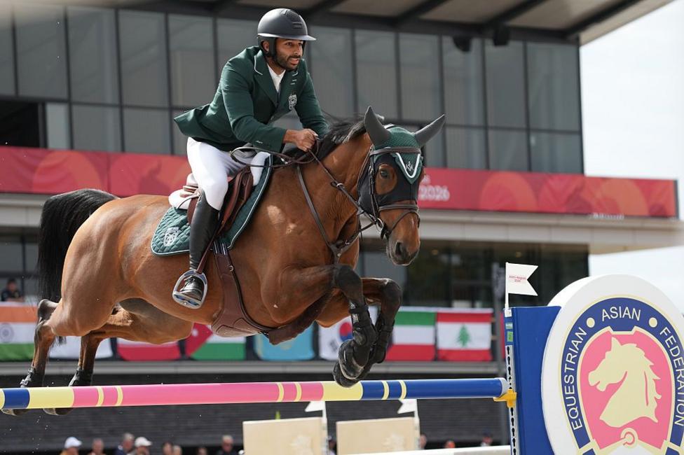 Saad Abdullah S Alajmi of Saudi Arabia and horse clear a jump during an equestrian event. Alajmi wears a dark green jacket, white breeches and a helmet, while the horse is fitted with protective leg wraps and a green ear bonnet.
