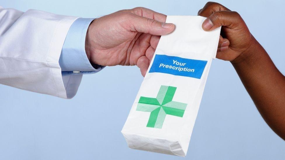 Бумажный пакет с надписью «your prescription» белым цветом внутри синей рамки. На белом бумажном пакете изображён зелёный крест.