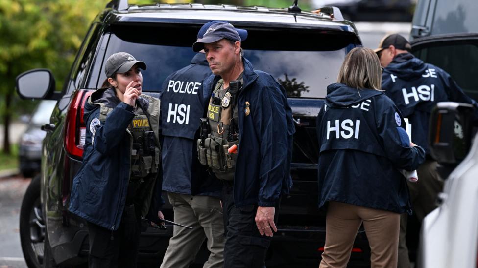 Men and women wearing blue jackets reading "Police HSI" stand behind a black parked car, two are turned toward the camera