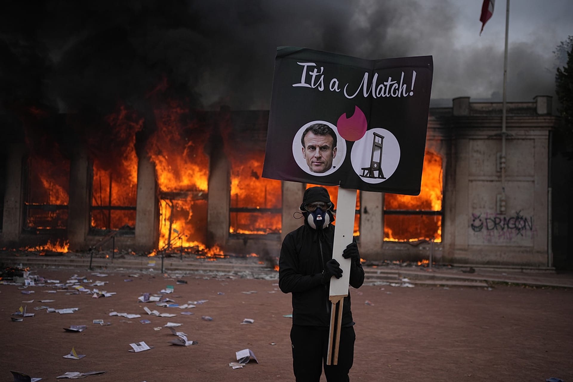 A protester holds a board with a photo of French president Emmanuel Macron in front of a burning tourism office during a protest in Lyon, France, Saturday, Oct. 17, 2026