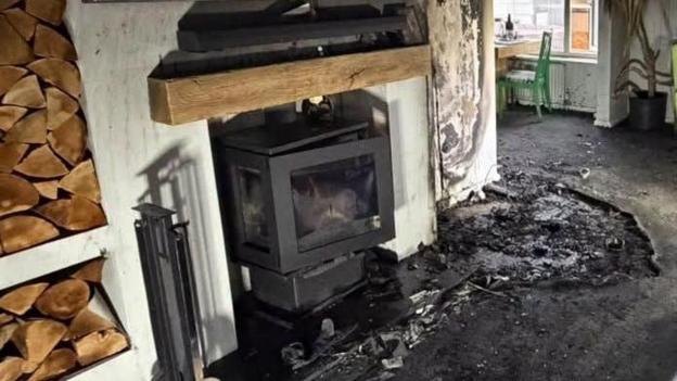 A log burner, in a room, with smoke and fire damage. Stacks of wood are to the left. The carpet is burnt and black. A table and green chair can be seen at the back of the room. A wood plinth is above the log burner, which is black.