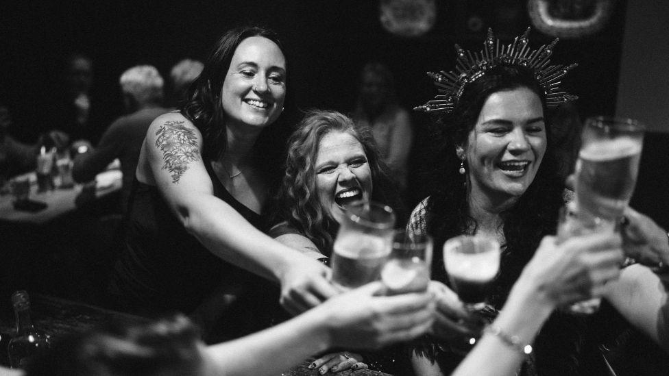 Black-and-white photo of several people gathered around a table, raising pint glasses in a toast. One woman wearing a decorative crown-like headpiece sits near the centre, while others lean in with outstretched arms holding drinks. The indoor setting is softly lit, with blurred figures and tables visible in the background.
