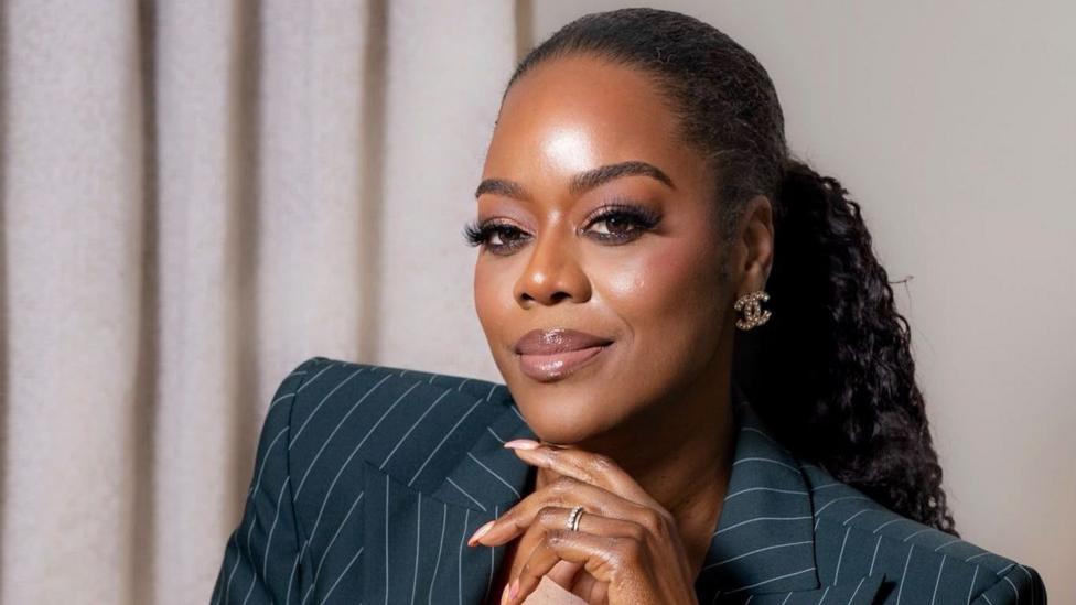 Antonia from the shoulders up. She is wearing a pin-striped navy suit and is holding her hand just under her chin. She has her long, black hair in a ponytail and is wearing earrings in the shape of the Chanel logo.