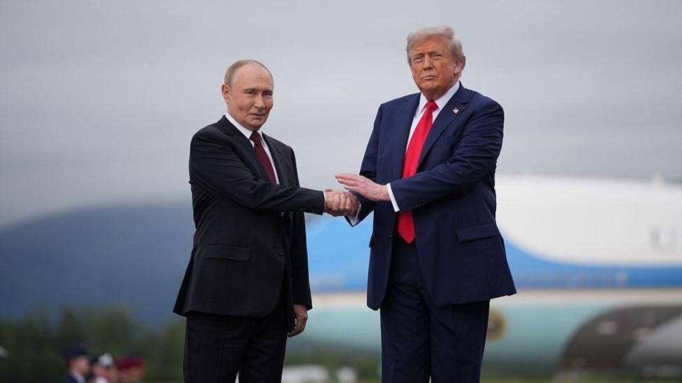 Two men shake hands as they pose for photographs. The shorter man, to the left, wears a black suit jacket and maroon tie, while the taller man, to the right, wears a dark blue suit jacket and red tie.