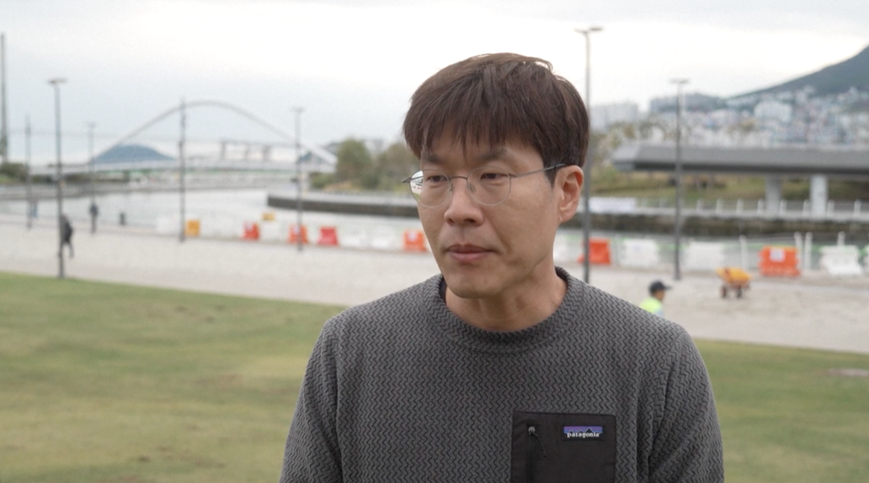 A person wearing glasses and a grey patterned Patagonia sweatshirt stands on a grassy area beside a waterfront canal in Busan, South Korea. In the background, a pedestrian bridge arches over the water, while construction barriers, lampposts and a riverside walkway line the canal. A hill with buildings is visible in the distance under an overcast sky. Visible text on the clothing reads