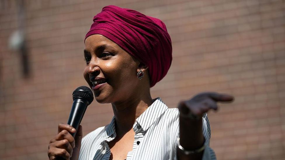Omar in magenta turban and black and white shirt with thin dark stripes, holds out her left hand while her right one holds a black microphone