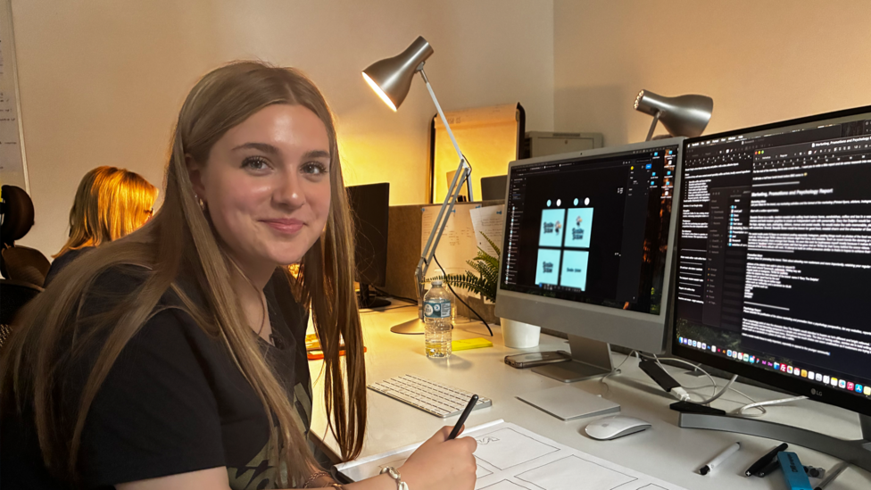 Jasmine is sitting in an office with two monitors in front of her and a large pad of drawing paper on her desk