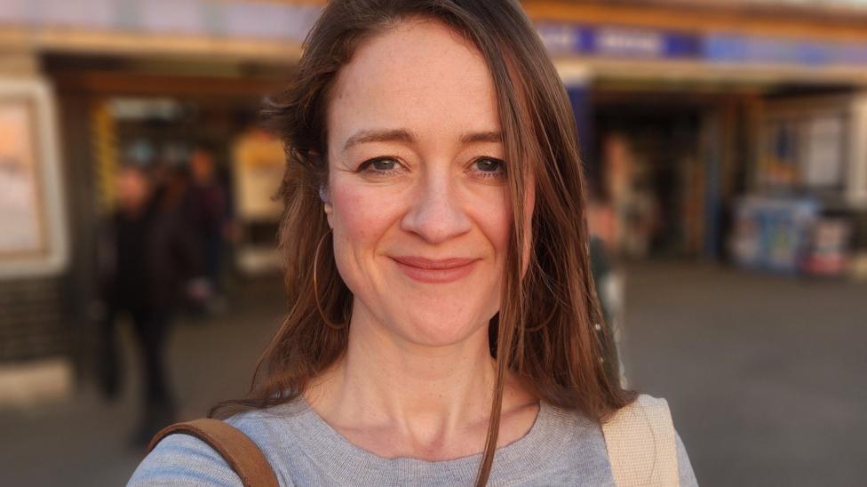Candice Gallagher with long brown hair and big hoop earrings smiles at the camera. A blurred London Underground station is seen behind her.