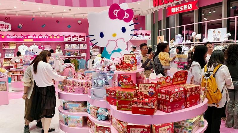 Ярко освещённый магазин товаров в тематике Hello Kitty, наполненный розовыми витринами и продукцией. В центре круглой витрины, уставленной игрушками, подарочными наборами и продукцией в коробках, стоит большая статуя Hello Kitty.