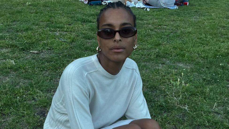 Summer wears a white jumper and sits on the grass. She has black hair braided back and wears sunglasses and gold earrings.