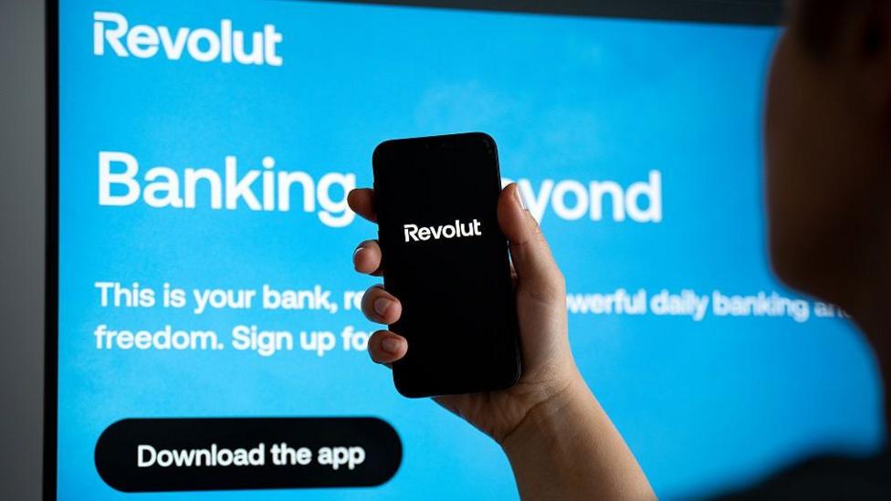 A person holding a smartphone displaying the Revolut app logo, in front of a display showing a bright blue Revolut advertisement.