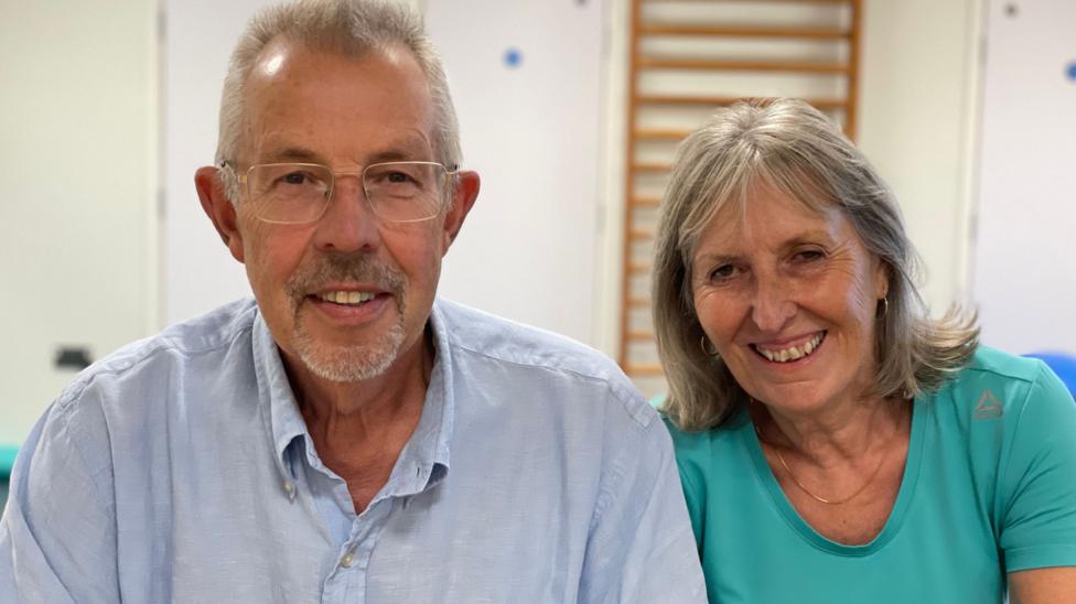 Steve and Angela Bucksey sit side-by-side smiling - Steve has grey hair and is wearing a light blue shirt with a collar, and Angela has shoulder-length greyish hair and wears a green round-necked T-shirt and a necklace