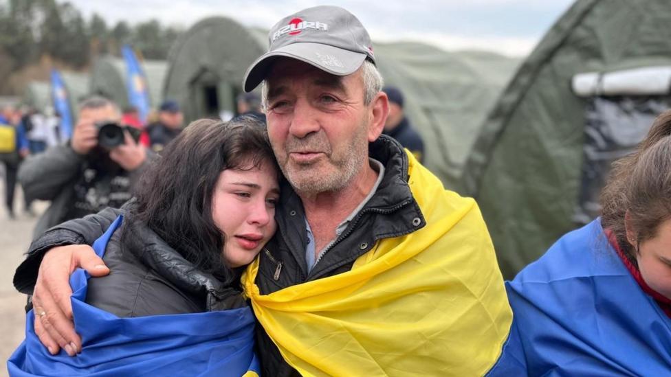 Kul rang kiyim kiygan Mixail köz yoşli qizlaridan birini qöltiğidan quçoqlab turibdi; qizining başi uning kökragiga qöyilgan. İkkalasi ham Ukraina bayroqlariga öralgan