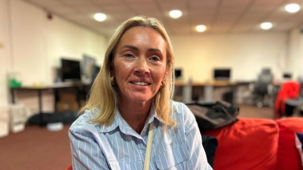 Tracey Rogerson with shoulder-length hair wearing a light blue striped shirt, seated indoors in a large room with desks, computer screens and red beanbags in the background.